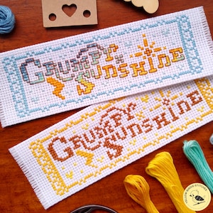 May include: Two cross-stitch bookmarks with the words "Grumpy Sunshine" in orange and yellow. One bookmark has a blue border, the other has a yellow border. The bookmarks are on a wooden surface with embroidery floss.