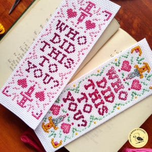 K&ouml;nnte beinhalten: Zwei Kreuzstich-Lesezeichen mit rosa und gr&uuml;nen Blumenmustern. Die Lesezeichen zeigen den Text "Who did this to you?" in einer fetten, dunkelrosa Schrift.