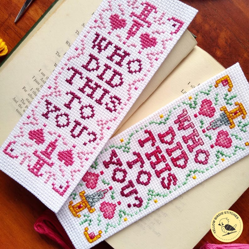 K&ouml;nnte beinhalten: Zwei Kreuzstich-Lesezeichen mit rosa und gr&uuml;nen Blumenmustern. Die Lesezeichen zeigen den Text "Who did this to you?" in einer fetten, dunkelrosa Schrift.