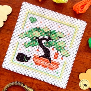 May include: Cross-stitch embroidery of a bonsai tree in a terracotta pot, with green foliage and small orange accents. A black cat rests near the pot. The design is framed with a green border on white fabric.
