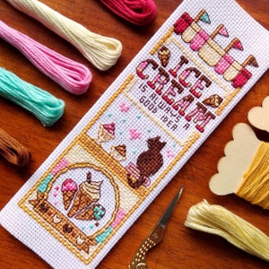 Peut inclure: Un marque-page brodé avec un thème de crème glacée. Le marque-page présente le texte "ICE CREAM IS ALWAYS A GOOD IDEA" et des illustrations de cornets de glace et d'un chat. La palette de couleurs comprend du rose, du marron et du crème, avec divers fils à broder en arrière-plan.