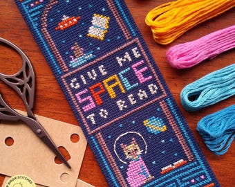 May include: A navy blue cross-stitch bookmark with the text "GIVE ME SPACE TO READ". The bookmark features a cat in a spacesuit, planets, and a UFO. Colorful embroidery floss and scissors are in the background.