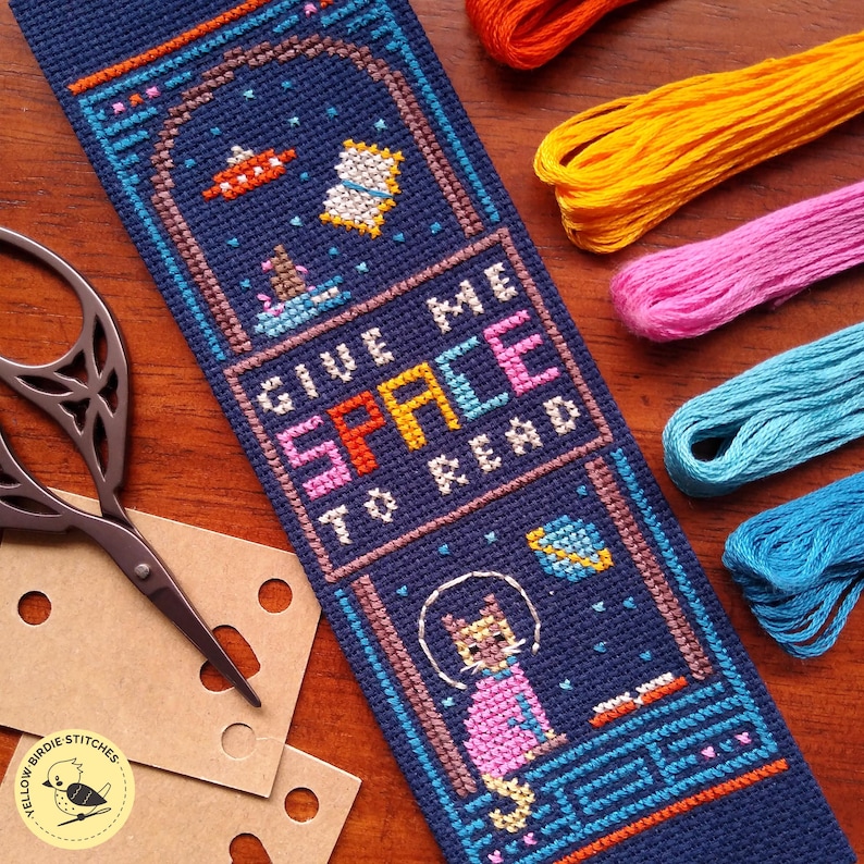 May include: A navy blue cross-stitch bookmark with the text "GIVE ME SPACE TO READ". The bookmark features a cat in a spacesuit, planets, and a UFO. Colorful embroidery floss and scissors are in the background.