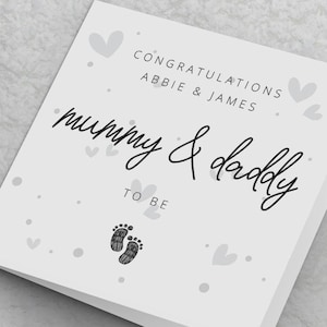 May include: A white card with black text that reads "Congratulations Abbie & James Mummy & Daddy To Be" with a small grey footprint graphic. The card is decorated with small grey hearts and dots.