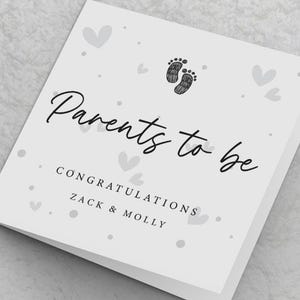 May include: White card with black text that reads "Parents to be" and "Congratulations Zack & Molly". The card is decorated with small grey hearts and a grey footprint graphic.
