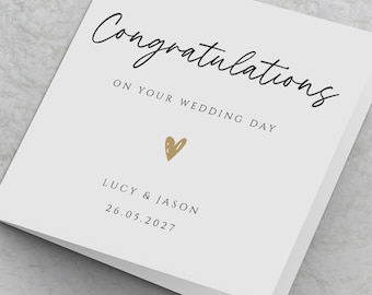Pode incluir: Um cartão de felicitações de casamento quadrado e branco com o texto em preto Congratulations ON YOUR WEDDING DAY, um pequeno ícone de coração dourado e os nomes Lucy & Jason com a data 26.05.2027 impressos na capa frontal.
