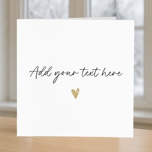 May include: A square white greeting card featuring the black cursive text Add your text here centered on the front. Below the text is a small, hand-drawn gold heart icon. The card is presented in a clean, minimalist style with a matte finish.