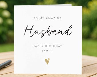 May include: A white greeting card with the words "TO MY AMAZING Husband HAPPY BIRTHDAY JAMES" in black script. A small gold heart is at the bottom. The card is on a wooden surface.