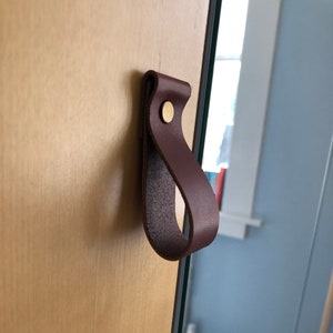 Puede incluir: Un tirador de armario de cuero marrón con un tornillo de latón. El tirador está fijado a una puerta de armario de madera.