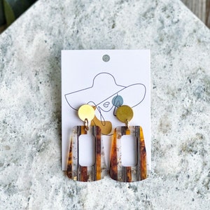 May include: A pair of gold and brown geometric earrings with a rectangular shape and a striped pattern. The earrings have a gold metal top and a clear resin base.