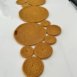Op de afbeelding: Mosterdgele gehaakte tafelloper en bijpassende onderzetters. De loper bestaat uit cirkelvormige stukken van verschillende grootte, en de onderzetters zijn ook cirkelvormig. Gemaakt van een getextureerd materiaal.