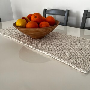 Könnte beinhalten: Ein gewebter, cremefarbener Tischläufer mit einem strukturierten Muster. Eine Holzschale mit Orangen und Zitronen steht auf dem Läufer. Der Tisch ist weiß, und im Hintergrund stehen dunkelgraue Stühle.