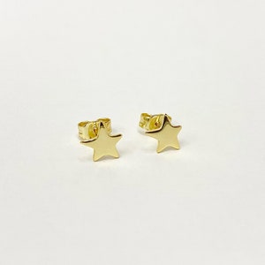 May include: A pair of small, gold-coloured star-shaped stud earrings. The earrings have a simple, five-pointed star design and are set against a white background. The earrings are likely made of a shiny metal.