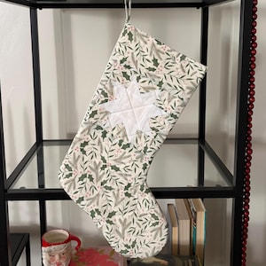 May include: A fabric Christmas stocking featuring a repeating pattern of green holly leaves, pine needles, and small pink berries on a cream background, accented with a white quilted star patch centered on the front of the stocking.