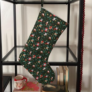 Puede incluir: Un calcetín navideño de color verde oscuro con un patrón repetitivo de perros pequeños con gorros de Papá Noel y bufandas, intercalados con pequeñas agujas de pino y bayas. El calcetín tiene un forro interior blanco y cuelga de un gancho negro.