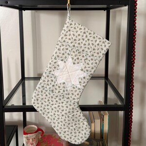 Puede incluir: Un calcetín navideño acolchado con un patrón floral en verde claro y blanco y un diseño de estrella blanca central. El calcetín tiene un lazo de tela en la parte superior para colgarlo y está construido con costuras visibles en la tela.