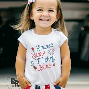 May include: A young child wearing a white t-shirt with the text "Stripes, Stars & Mickey Bars" in red and blue. The child is also wearing red, white, and blue striped shorts and a bow in their hair. The child is smiling.