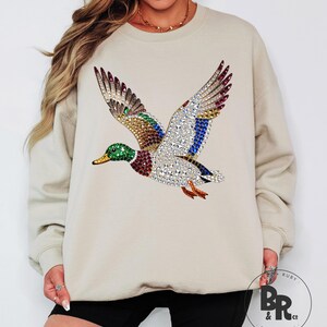 May include: Beige sweatshirt featuring a detailed, bejeweled duck in flight. The duck design incorporates various colors, including green, red, blue, and silver, creating a sparkling effect. The sweatshirt has a crew neck and long sleeves.