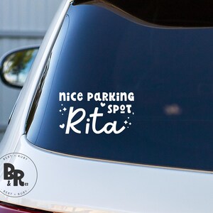 Puede incluir: Adhesivo de vinilo blanco para coche con el texto "nice parking spot, Rita" en una fuente juguetona. El diseño incluye pequeños corazones y detalles brillantes. El adhesivo está en la ventana trasera de un coche blanco.