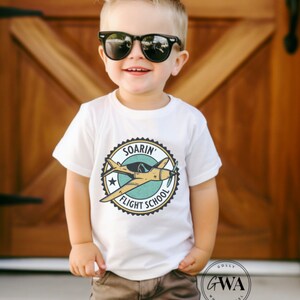 May include: A white t-shirt with a graphic of a yellow airplane in a blue and green circle with the text "Soarin' Flight School".