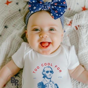 May include: A white short-sleeved bodysuit featuring a graphic of George Washington wearing sunglasses with the text TOO COOL FOR BRITISH RULE and EST 1776. The outfit includes a blue headband with a large bow patterned with red and white stars.
