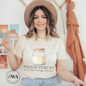 May include: A woman wearing a light beige t-shirt with the text "Proud Parent of a Sourdough Starter" and a drawing of a glass jar with a sourdough starter inside. The woman is wearing a brown hat and has her hair down. She is holding up two fingers in a peace sign.