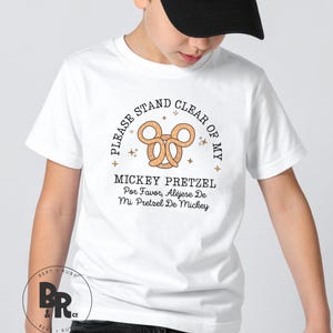 May include: White t-shirt with a cartoon pretzel design and the text "PLEASE STAND CLEAR OF MY MICKEY PRETZEL". The design includes a pretzel, stars, and text in English and Spanish. The shirt is for children.