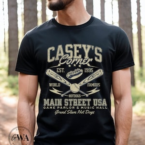 May include: Black t-shirt with a vintage-style graphic design featuring the text "Casey's Corner" with baseball bats, a baseball, and hot dogs. The text "EST. 1995" is also included. The graphic also includes the text "WORLD FAMOUS HOTDOGS MAIN STREET USA GAME PARLOR & MUSIC HALL Grand Slam Hot Dogs".