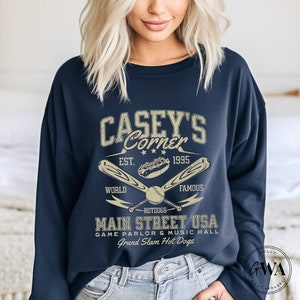 May include: A navy blue sweatshirt with a vintage-style graphic design featuring the text "Casey's Corner" with a baseball bat and ball, hot dog, and the text "Est. 1995" and "World Famous". The graphic also includes the text "Main Street USA, Game Parlor & Music Hall, Grand Slam Hot Dogs".
