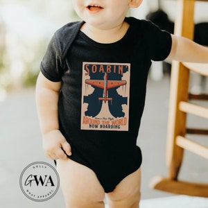 May include: A black baby onesie with a vintage travel poster design featuring a red airplane flying through clouds. The poster says "Soarin' Around the World Now Boarding".