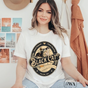 May include: A white t-shirt with a black and gold circular graphic featuring a black cat, a skull, and a candle. The graphic reads "Hand Dipped Clean Burning Black Cat Candles by Thackery Binx Since 1893 Salem, MA".