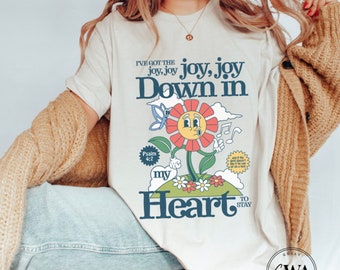 May include: A white t-shirt with a graphic design of a cartoon flower with a smiling face. The flower has red petals and a yellow center. The text "I've got the joy, joy, joy, joy Down in my Heart to stay" is printed on the shirt. The text "Psalm 4:7" is also printed on the shirt.