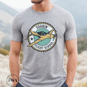 May include: A gray t-shirt with a vintage-style graphic of a yellow airplane flying in a circle with the text "Soarin' Flight School".