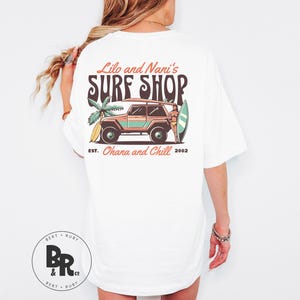 May include: White t-shirt with a colorful graphic on the back. The graphic features a vintage-style surf shop design with a jeep, palm tree, and surfboard. Text includes "Lilo and Nani's SURF SHOP", "EST. Ohana and Chill 2002".
