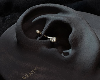 Könnte beinhalten: Ein goldfarbener Helix-Ohrring mit einem tropfenförmigen, klaren Edelstein. Der Ohrring wird auf einem dunkelgrauen, ohrförmigen Display präsentiert. Das Wort "BEATTI" ist auf dem Display geprägt.