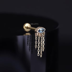 May include: Gold body jewellery with a blue marquise gemstone and a chain dangle. The jewellery is on a black background. BEATTI is written on the jewellery.