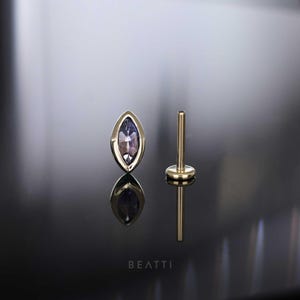 Peut inclure: Boucle d'oreille à tige en or en forme de marquise avec une pierre précieuse violette, à côté d'une tige en or assortie. L'image est sur une surface noire réfléchissante. Le mot "BEATTI" est en bas.