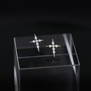 May include: Two cross-shaped stud earrings. One is silver-toned, the other gold-toned. Both feature clear, round gemstones. The earrings are displayed on a clear, rectangular stand against a black background.