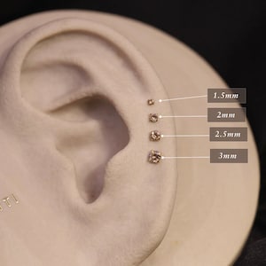 May include: A close-up of a model ear with five different sized clear gemstone earrings. The earrings are labeled with their size in millimeters: 1.5mm, 2mm, 2.5mm, and 3mm.