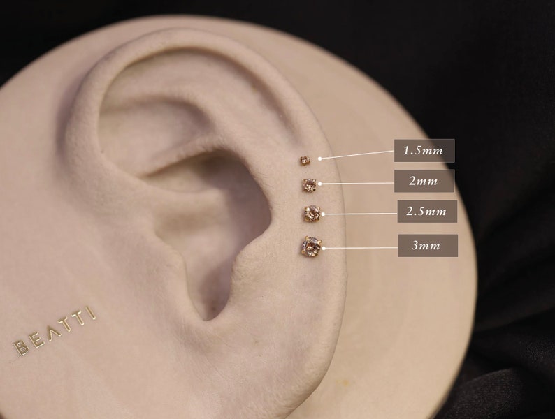May include: A close-up of a model ear with five different sized clear gemstone earrings. The earrings are labeled with their size in millimeters: 1.5mm, 2mm, 2.5mm, and 3mm.