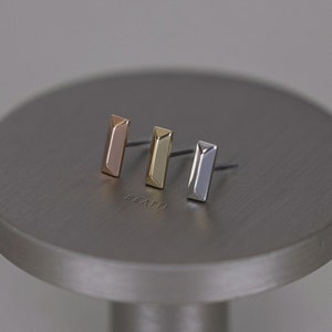 May include: Three stud earrings in rose gold, gold, and silver, each with a rectangular bar design. The earrings are displayed on a gray, circular surface. The word "BEATTI" is subtly engraved on the surface.