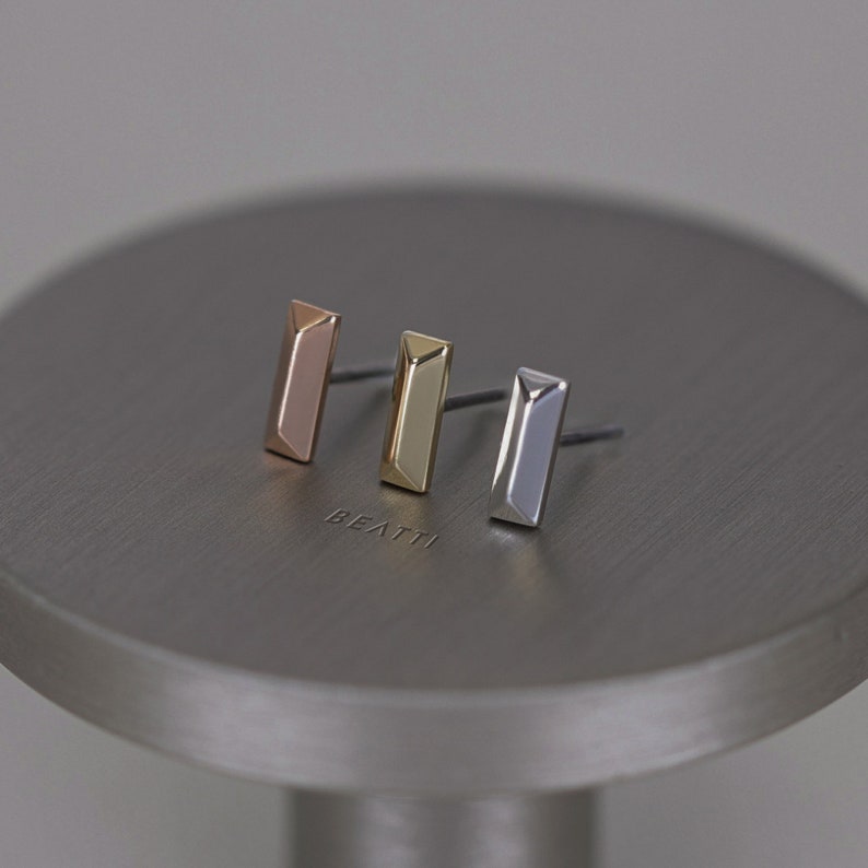 May include: Three stud earrings in rose gold, gold, and silver, each with a rectangular bar design. The earrings are displayed on a gray, circular surface. The word "BEATTI" is subtly engraved on the surface.