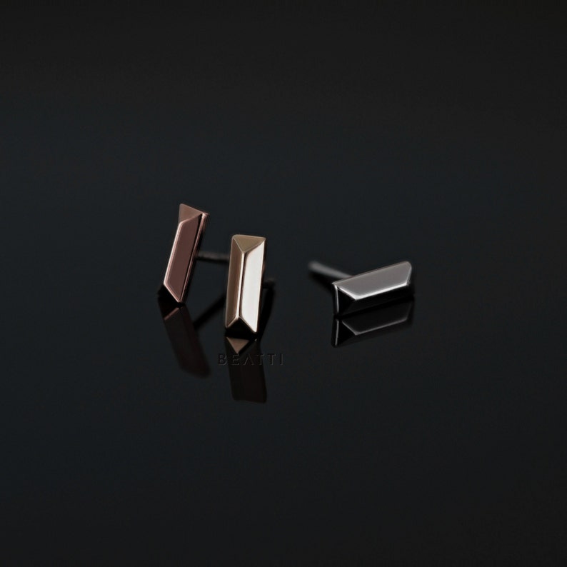 May include: Three pairs of geometric bar stud earrings in rose gold, gold, and silver tones. The earrings have a minimalist design with a rectangular shape and angled edges. The earrings are displayed on a reflective black surface.