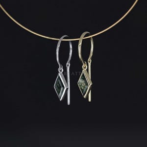 May include: A pair of dangle earrings, one silver and one gold, each featuring a diamond-shaped green stone. The earrings hang from a thin gold wire against a black background. The earrings have a modern, minimalist design.