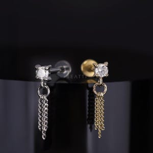 May include: Two stud earrings with a dangling chain design. One earring is silver-toned with a silver chain, and the other is gold-toned with a gold chain. Each earring features a clear, round gemstone.