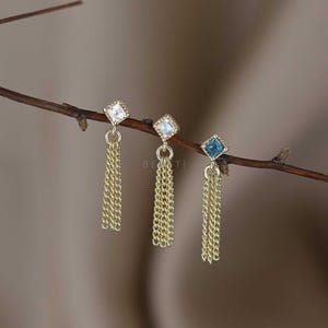 May include: Three gold-coloured dangle earrings with square-cut gemstones in clear, iridescent, and blue hues. Each earring features a chain tassel design. The earrings are displayed on a thin, brown branch against a blurred background.