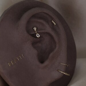 May include: Close-up of an ear with multiple gold earrings. A teardrop-shaped earring hangs from the upper ear, and a small bar earring is visible. The word "BEATTI" is printed in gold on the ear.