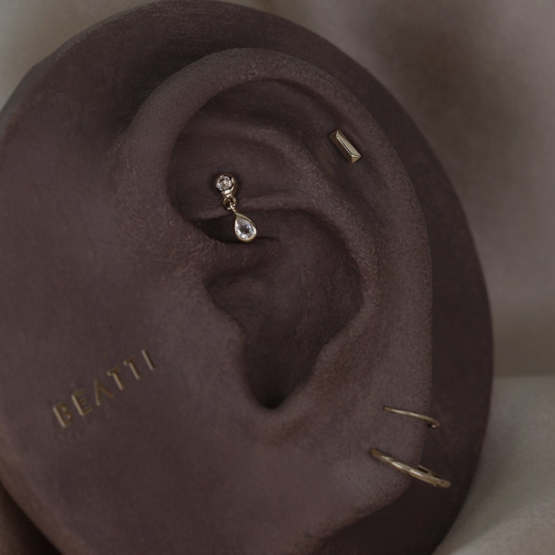 May include: Close-up of an ear with multiple gold earrings. A teardrop-shaped earring hangs from the upper ear, and a small bar earring is visible. The word "BEATTI" is printed in gold on the ear.