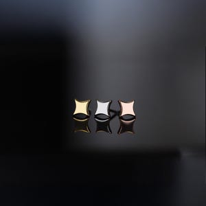Peut inclure: Trois boucles d'oreilles à clous en or, argent et or rose. Chaque boucle d'oreille a une forme carrée aux côtés incurvés. Les boucles d'oreilles sont présentées sur une surface noire réfléchissante.