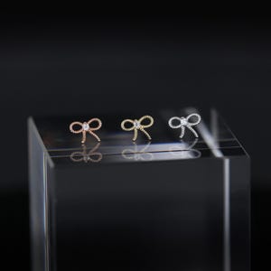 May include: Three small bow-shaped earrings in rose gold, gold, and silver, each with a small, clear gemstone in the center. The earrings are displayed on a clear, rectangular block against a dark background.